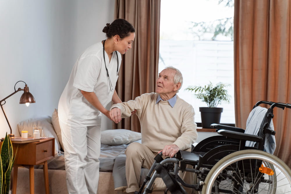 Une infirmière aide un senior à s'asseoir dans un fauteuil roulant. L'ambiance est calme et rassurante, comme dans une maison de retraite à Vincennes.