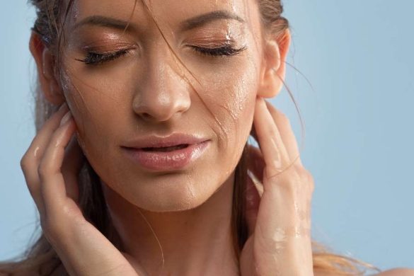 Jeune femme les yeux fermés, le visage mouillé et les mains sur les joues, en train de nettoyer son visage à l'eau sur fond bleu clair.